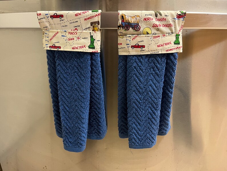 May include: Two blue kitchen towels with a white and yellow patterned fabric border. The border features a map of the United States with various state names and landmarks. One towel has a red truck and the other has a covered wagon.