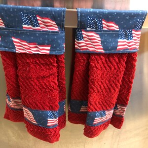 May include: Two red kitchen towels with a blue and white American flag print on the top and bottom. The towels are hanging on a silver towel rack.