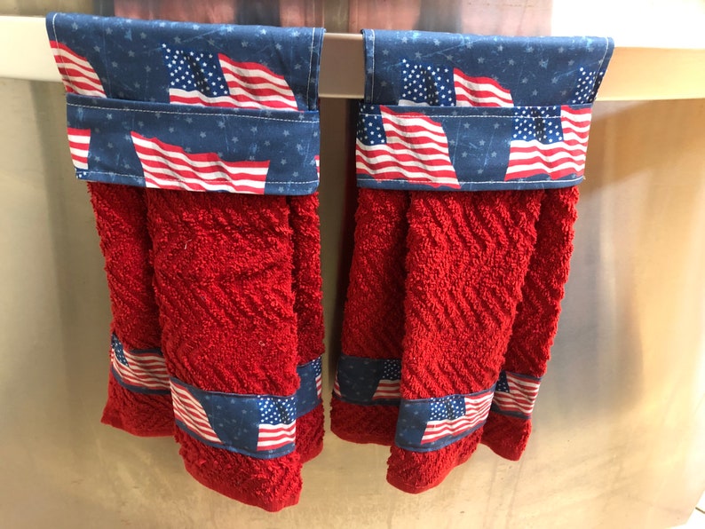 May include: Two red kitchen towels with a blue and white American flag print on the top and bottom. The towels are hanging on a silver towel rack.