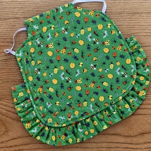 May include: A green apron featuring a repeating pattern of various insects including bees, beetles, and spiders in yellow, orange, blue, and black. The apron has a ruffled hem along the bottom and sides, and includes white elastic straps for securing.