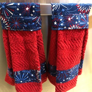 May include: Two red kitchen towels with a blue and white firework print border. The towels are hanging on a silver towel rack.