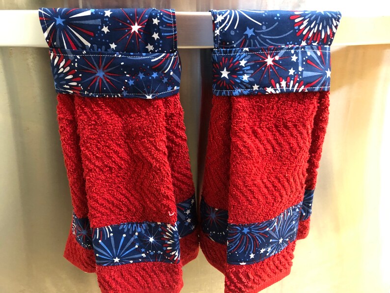 May include: Two red kitchen towels with a blue and white firework print border. The towels are hanging on a silver towel rack.