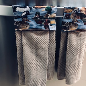 May include: Two brown kitchen towels with a duck and bird print border hanging on a silver towel rack.