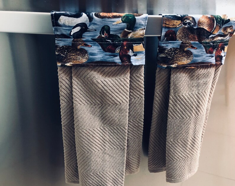 May include: Two brown kitchen towels with a duck and bird print border hanging on a silver towel rack.