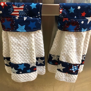 May include: Two white kitchen towels with blue and red star and firework patterned fabric accents. The towels are hanging on a silver rack.