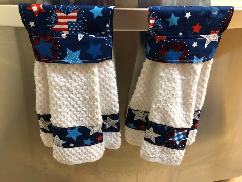 May include: Two white kitchen towels with blue and red star and firework patterned fabric accents. The towels are hanging on a silver rack.