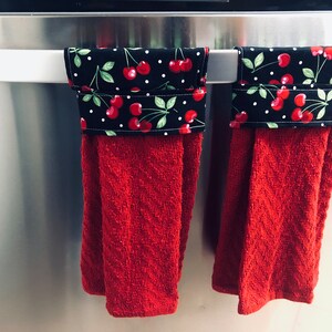 May include: Two red kitchen towels with black and white cherry patterned fabric accents hanging on a silver refrigerator handle.