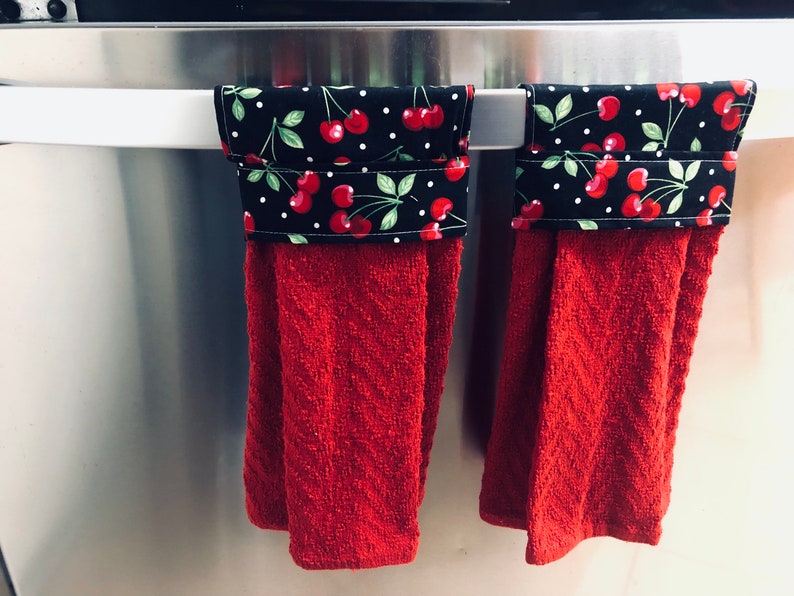 May include: Two red kitchen towels with black and white cherry patterned fabric accents hanging on a silver refrigerator handle.
