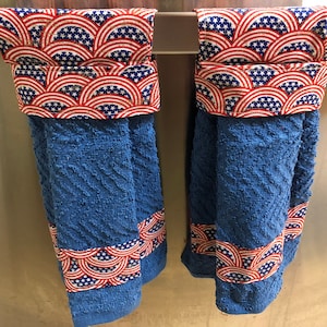 May include: Two blue kitchen towels with red, white and blue patterned fabric accents. The towels are hanging on a silver towel rack.