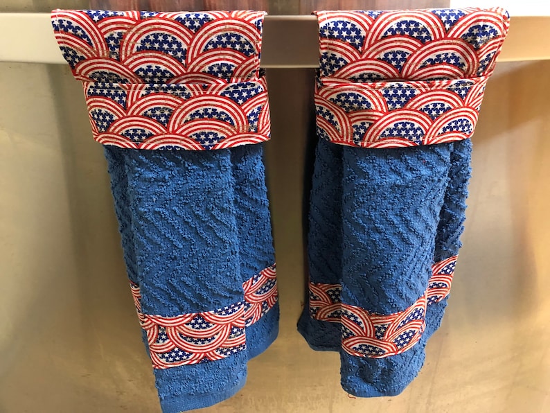 May include: Two blue kitchen towels with red, white and blue patterned fabric accents. The towels are hanging on a silver towel rack.
