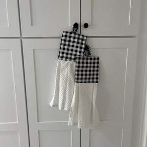 May include: Two white kitchen towels with black and white checkered fabric accents. The towels are hanging on a white cabinet door.