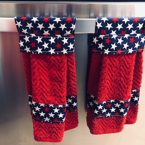 May include: Two red kitchen towels with white stars on a blue background. The towels are hanging on a silver rack.