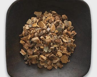 May include: A dark brown, square wooden bowl filled with a pile of dried, irregularly shaped pieces. The pieces vary in color, with shades of brown, tan, and hints of gray and green. The bowl's edges are rounded, and the wood grain is visible.