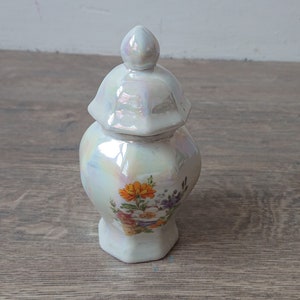 Puede incluir: Un pequeño tarro de porcelana blanca con tapa, decorado con un patrón floral en tonos naranja, rosa y morado. El tarro tiene un acabado perlado.