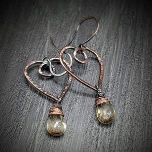 May include: A pair of handcrafted copper earrings. Each earring features a heart-shaped frame with a wire-wrapped, teardrop-shaped, faceted, light-yellow gemstone. The ear wires are a darker metal.