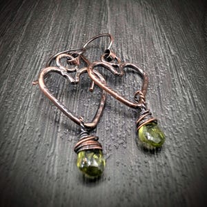 May include: Copper heart-shaped earrings with a textured finish. Each earring features a wire-wrapped, olive-green gemstone pendant. The earrings are displayed on a dark, textured surface.