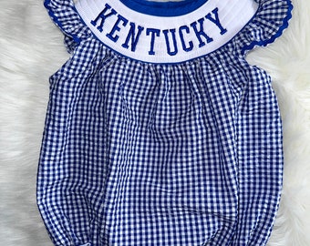 May include: Blue and white gingham baby onesie with a white ruffle trim and a blue and white collar with the word "KENTUCKY" embroidered in white.