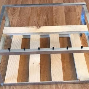 Puede incluir: Un marco de metal plateado con listones de madera en el interior. El marco es rectangular y tiene un diseño sencillo. Los listones de madera están dispuestos horizontalmente y están fijados al marco con tornillos.