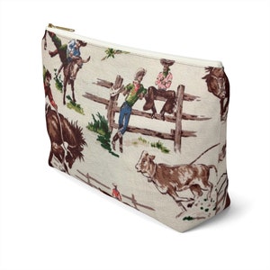 Puede incluir: Una bolsa blanca con cremallera con un estampado vintage de vaquero que presenta vaqueros montando caballos y toros, y una valla de madera. El estampado es en tonos de marr&oacute;n, verde, rojo y azul.