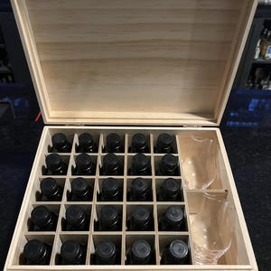 May include: A light-colored wooden box with a hinged lid, containing twenty-five square compartments. Twenty-three compartments hold small dark glass bottles with black screw caps, and two adjacent compartments contain clear glass tasting glasses.