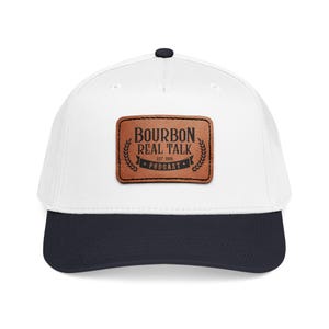 May include: A white baseball cap with a dark navy blue brim and a rectangular brown leather patch centered on the front. The patch features the black text BOURBON REAL TALK, EST. 2020, PODCAST, surrounded by decorative wheat-like leaf patterns.