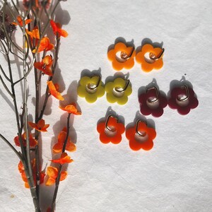 May include: Six pairs of flower-shaped earrings in various colors, including orange, yellow, and burgundy. The earrings are made of resin and have a gold hoop.