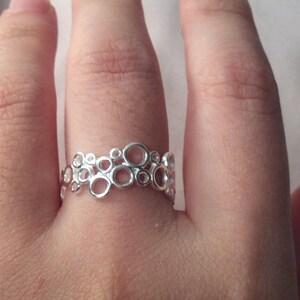 Puede incluir: Un anillo plateado con un dise&ntilde;o calado &uacute;nico con c&iacute;rculos interconectados. El anillo se lleva en un dedo, mostrando su intrincado patr&oacute;n y superficie reflectante. La joya es un accesorio elegante.