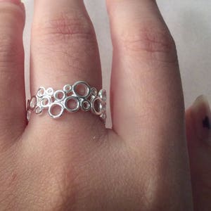 Puede incluir: Un anillo plateado con un dise&ntilde;o de burbujas. El anillo presenta m&uacute;ltiples c&iacute;rculos interconectados de varios tama&ntilde;os, creando una est&eacute;tica abierta y aireada. El anillo se lleva en un dedo, mostrando sus detalles delicados e intrincados. El anillo est&aacute; hecho de un metal brillante y reflectante.