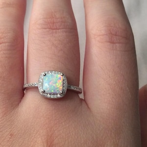 May include: A silver ring with a square-shaped opal gemstone surrounded by smaller clear gemstones.