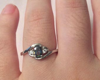 May include: A silver ring with a dark oval-shaped gemstone and two smaller clear gemstones on the band.