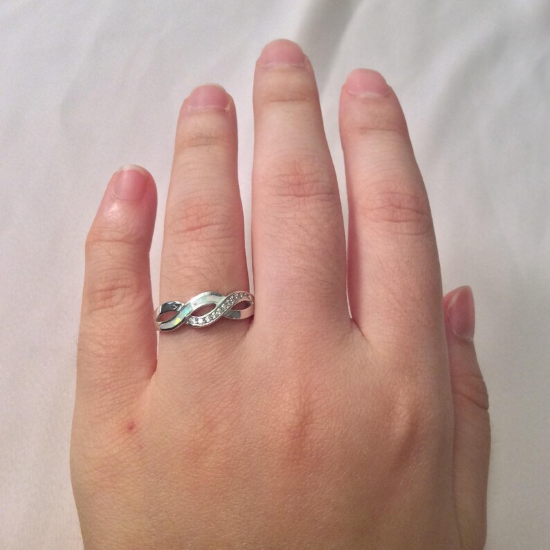May include: A silver-colored ring with an infinity symbol design. The ring features a series of intertwined loops, with small, clear gemstones set along one of the loops. The ring is displayed on a hand.