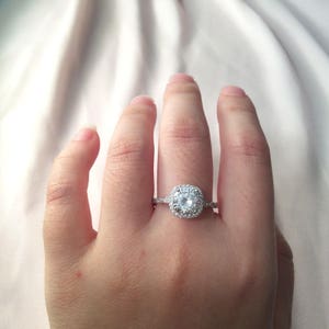 May include: A silver ring with a large, round, clear center stone surrounded by smaller stones. The ring is on a person's finger.