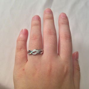 May include: A silver-colored ring with an infinity symbol design. The ring features a row of small, clear stones and an iridescent stone. The ring is on a hand.