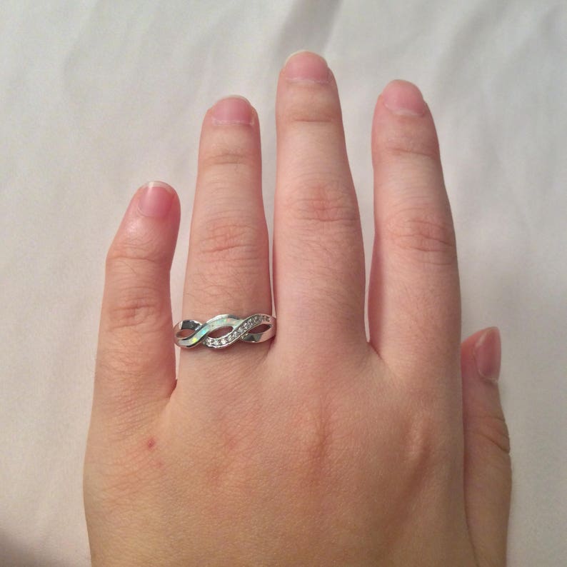 May include: A silver-colored ring with an infinity symbol design. The ring features a row of small, clear stones and an iridescent stone. The ring is on a hand.