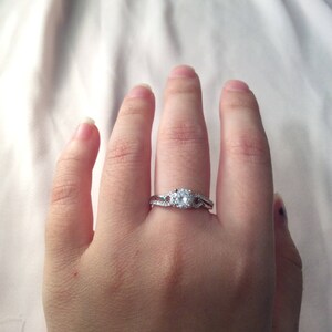 Op de afbeelding: Een zilveren ring met een grote, ronde, heldere edelsteen als middelpunt. De band heeft kleinere, heldere edelstenen en een gesplitst ontwerp. De ring wordt aan een vinger gedragen.