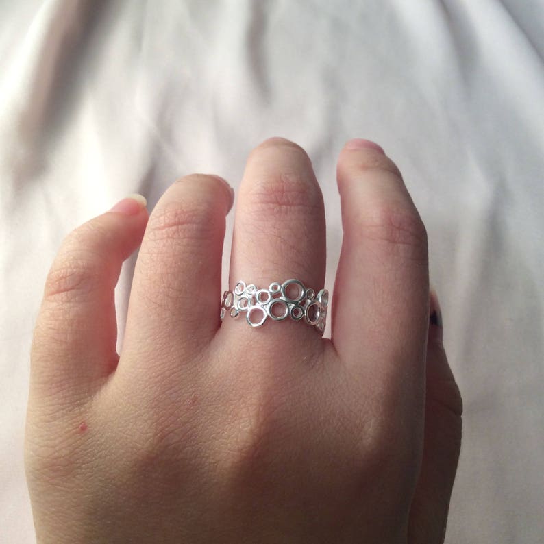 Puede incluir: Un anillo plateado con un dise&ntilde;o de c&iacute;rculos interconectados. El anillo se lleva en un dedo, mostrando su intrincado patr&oacute;n calado. El color plateado brillante del anillo contrasta con el tono de la piel, resaltando su delicado dise&ntilde;o. El anillo es una joya.
