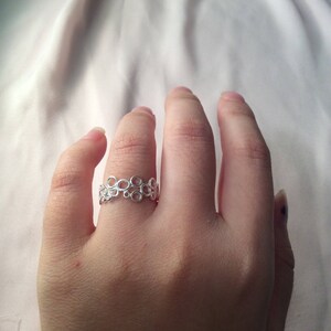 Puede incluir: Un anillo plateado con un delicado dise&ntilde;o calado de c&iacute;rculos interconectados. El anillo se lleva en un dedo, mostrando su intrincado patr&oacute;n y superficie reflectante. El dise&ntilde;o del anillo es una banda continua de peque&ntilde;as formas circulares.