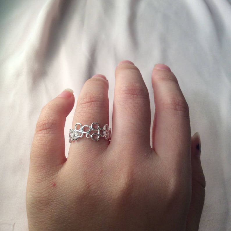 Puede incluir: Un anillo plateado con un delicado dise&ntilde;o calado de c&iacute;rculos interconectados. El anillo se lleva en un dedo, mostrando su intrincado patr&oacute;n y superficie reflectante. El dise&ntilde;o del anillo es una banda continua de peque&ntilde;as formas circulares.