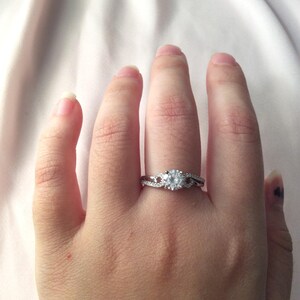 Op de afbeelding: Een zilveren ring met een grote, ronde, heldere edelsteen als middelpunt. De band heeft een gedraaid ontwerp met kleinere, heldere stenen. De ring zit aan een hand, tegen een zachte, lichtroze achtergrond.
