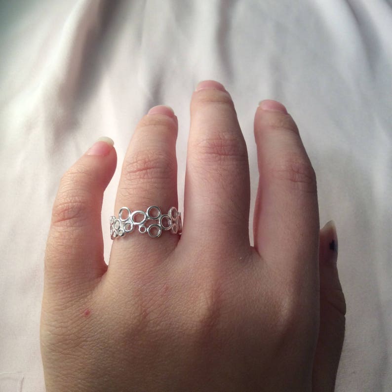 Puede incluir: Un anillo plateado con un dise&ntilde;o de burbujas se lleva en un dedo. El anillo presenta m&uacute;ltiples c&iacute;rculos peque&ntilde;os interconectados, creando una est&eacute;tica &uacute;nica y aireada. El anillo se encuentra sobre un fondo blanco suave.