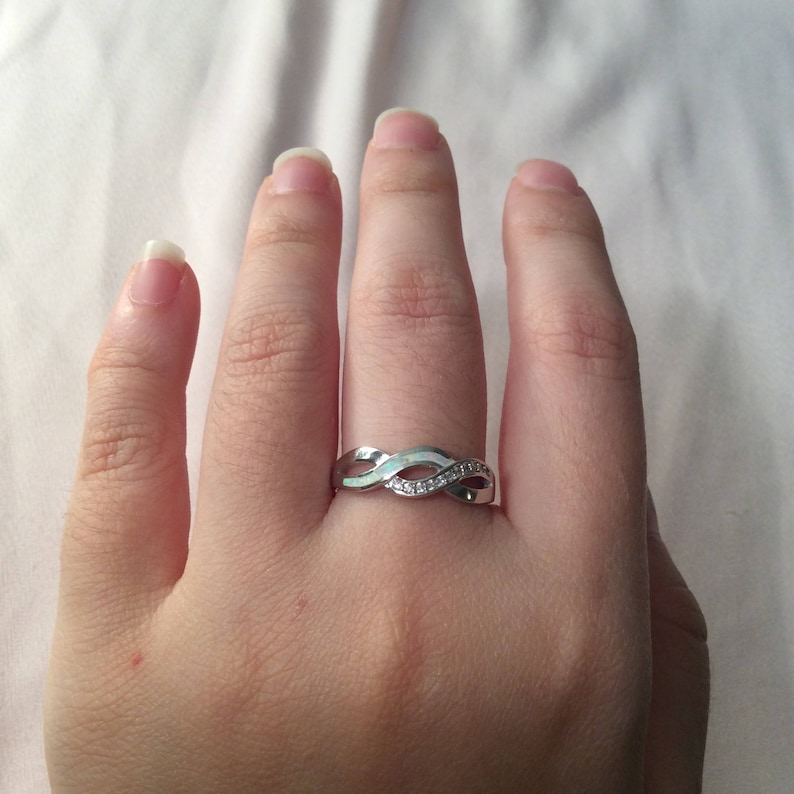 May include: A silver-colored ring with an infinity symbol design. The ring features a series of interwoven bands, with small, clear stones set along one section. The ring is worn on a finger.