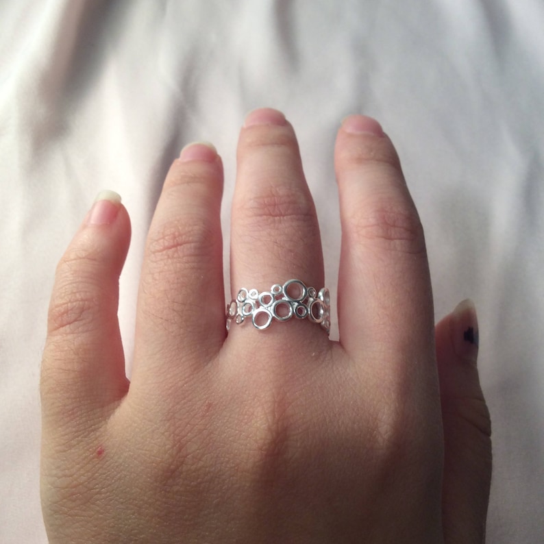 Puede incluir: Un anillo plateado con un dise&ntilde;o calado &uacute;nico con c&iacute;rculos interconectados. El anillo se lleva en un dedo, mostrando su intrincado patr&oacute;n y superficie reflectante. La joya es un accesorio elegante.