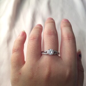 Op de afbeelding: Een zilveren ring met een grote, ronde, heldere edelsteen als middelpunt. De band heeft een gesplitst ontwerp met kleinere, heldere stenen. De ring wordt aan een vinger gedragen en toont zijn ingewikkelde details en schittering.
