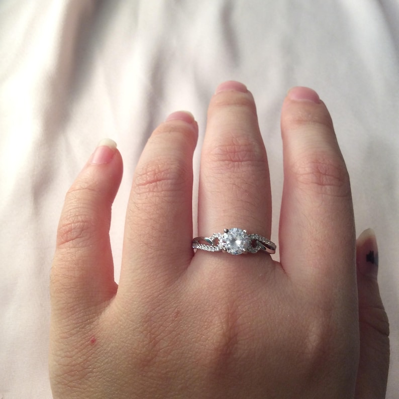Op de afbeelding: Een zilveren ring met een grote, ronde, heldere edelsteen als middelpunt. De band heeft een gesplitst ontwerp met kleinere, heldere stenen. De ring wordt aan een vinger gedragen en toont zijn ingewikkelde details en schittering.
