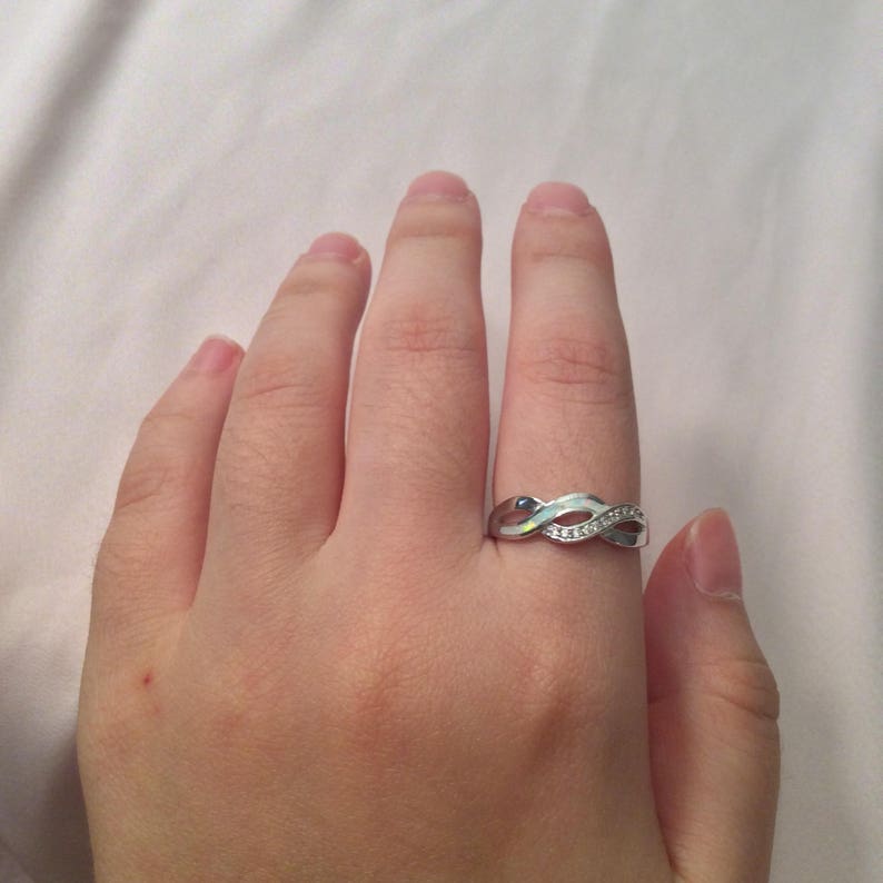May include: A silver-colored ring with an infinity symbol design. The ring features a series of small, clear stones set along one side, adding a touch of sparkle. The ring is worn on a finger, showcasing its elegant and timeless style.