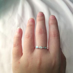 May include: A silver ring with three oval-shaped opals. The opals have a shimmering, iridescent quality, displaying a range of colors. The ring is set on a hand against a white background.