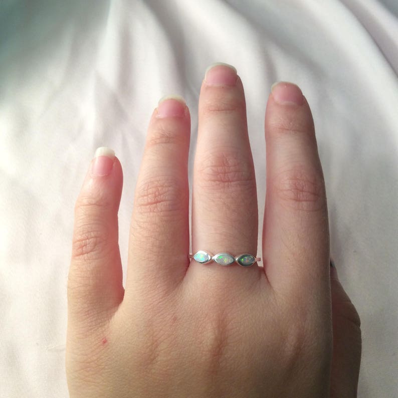 May include: A silver ring with three oval-shaped opals. The opals have a shimmering, iridescent quality, displaying a range of colors. The ring is set on a hand against a white background.