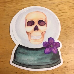 May include: A sticker featuring a watercolor illustration of a skull inside a snow globe. The skull is beige with dark purple eye sockets. A purple flower is on the base of the snow globe, which is dark green.