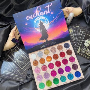 May include: A colorful eyeshadow palette with the word "enchant" and a witch silhouette. The palette features a variety of glittery shades, with names like "moon," "stars," and "magic." The brand name "CREATIVE | SOUL" is also visible.