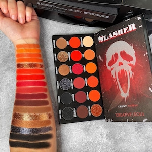 Può includere: Una palette di ombretti nera aperta con 15 tonalità in arancione, rosso, marrone e nero. La palette è accanto a un libro con la scritta "SLASHER" e una grafica a teschio. Campioni di ombretto su un braccio.